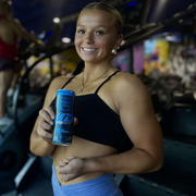 A smiling woman in workout attire holding a can of UD Energy Blue Raspberry flavor in a gym setting. The can's design, featuring a bulldog and the slogan "FEED YOUR BEAST," is visible as she poses, suggesting an active lifestyle complemented by the energy drink.